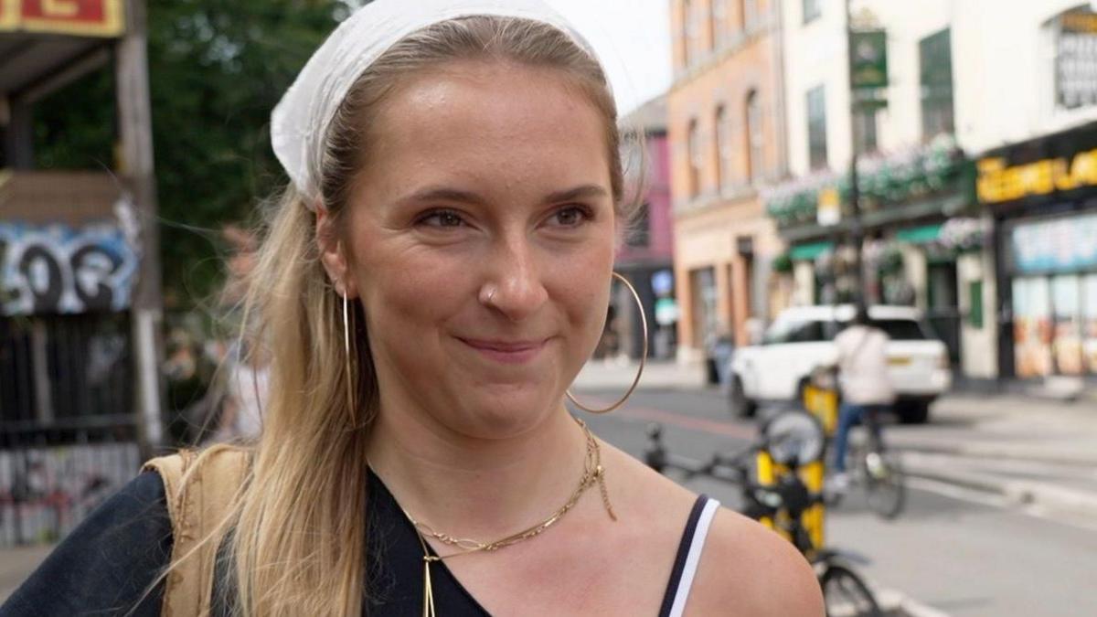 A white woman with long blond hair tied back by a white bandana. She has big hoop earrings and wears a black off the shoulder top and gold necklaces.
