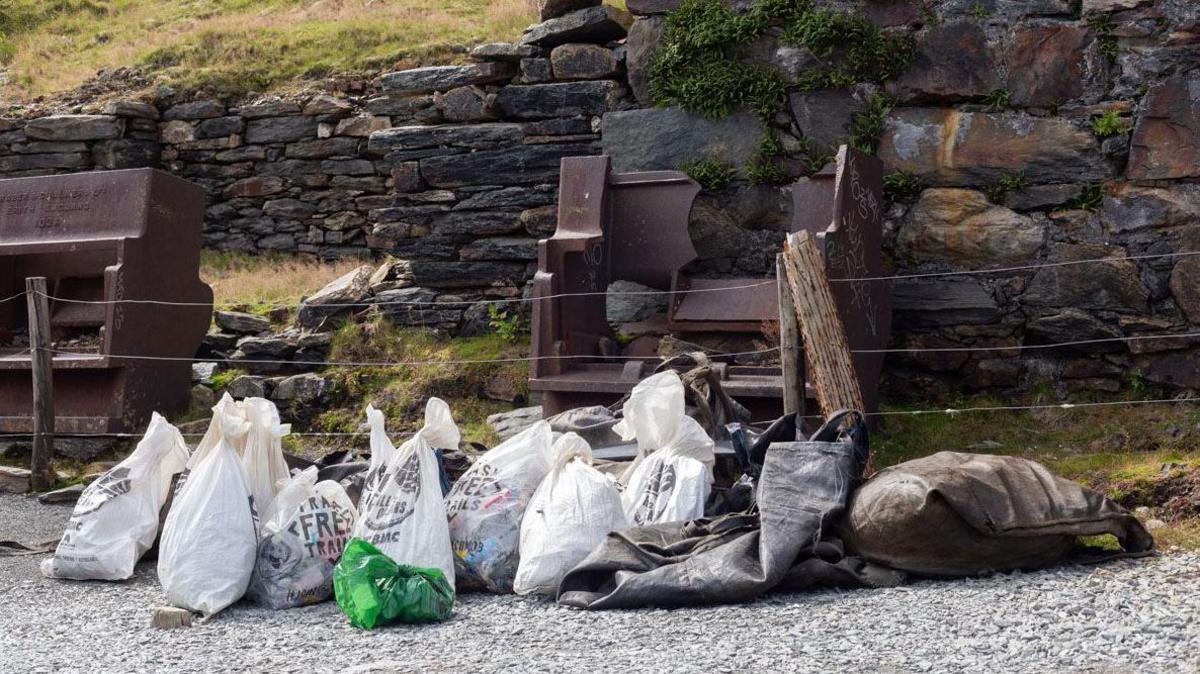 Several white plastic bags full of rubbish, piled on a stoney path with greenery behind.