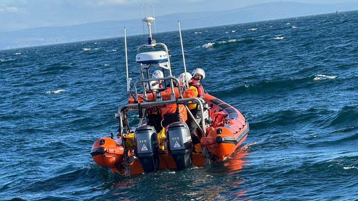 Orange life boat with crew wearing helmets on board seen in the sea from behind with engines at the back