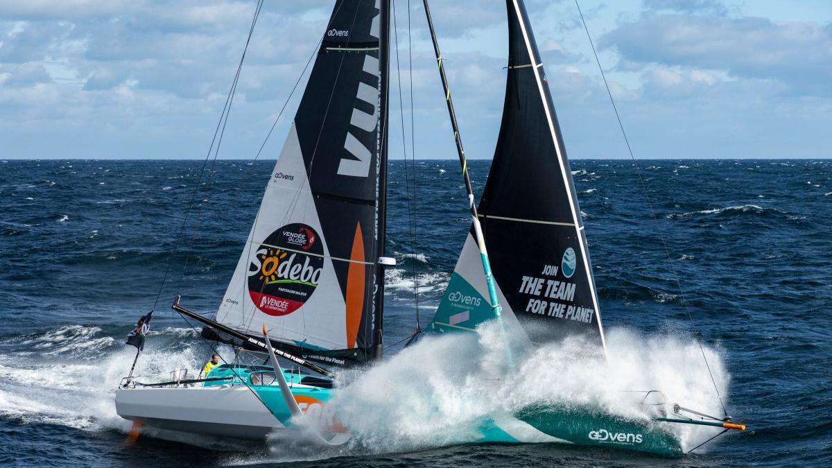 A sailaing boat going fast with spray with the ocean around it and a man in a  yellow sailing outfit in the cockpit.