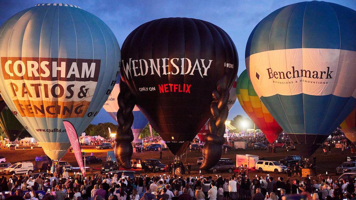 A black Wednesday Addams-themed hot air balloon is inflated on the ground next to a selection of other hot air balloons, at night. A large crowd looks up at the colourful balloons.
