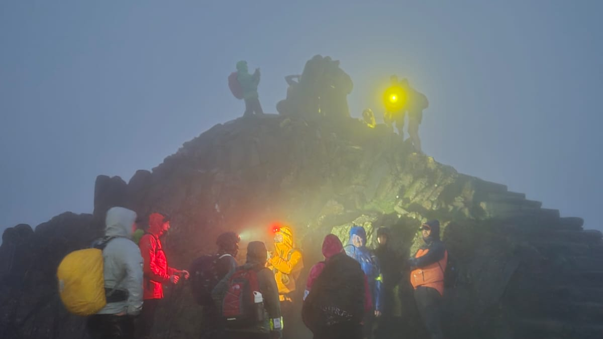The summit of Yr Wyddfa. the sky is dark and foggy There are people standing up on a lookout with a yellow torch and are taking photos of each other. Below them, down some stone steps, is another group of people in hiking gear, waterproof coats and head torches