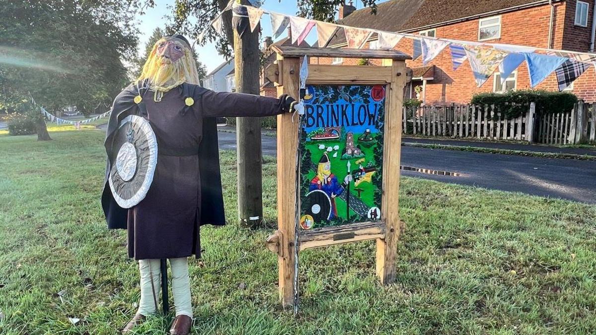 A scarecrow dressed in a brown outfit with white trousers and a cape. It is holding a shield and has a blonde beard. The scarecrow is clutching onto a wooden sign that says 'Brinklow'. Bunting lines the streets. 