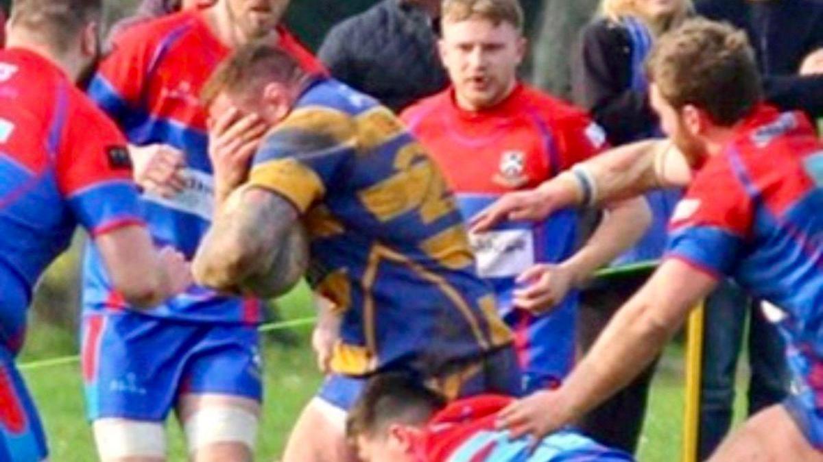 Ross yn ystod gêm rygbi, yng nghanol nifer o chwaraewyr eraill - mae'r bêl yn ei law chwith ac mae'n gafael yn ei wyneb gyda'r law dde 