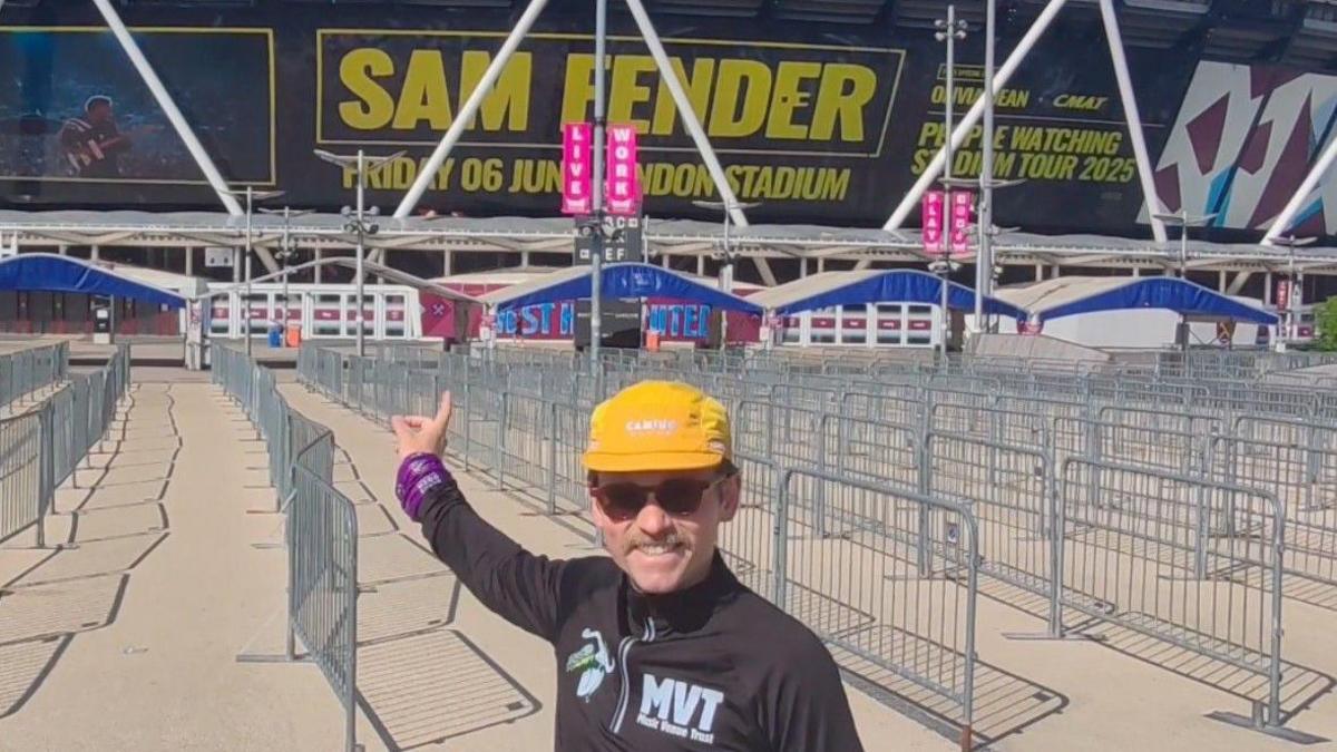 Andy Hobson, dressed in a running top and a yellow hat, is pictured standing outside the London Stadium. The stadium side reads "Sam Fender" in large yellow letters, as well as carrying the date of his performance.