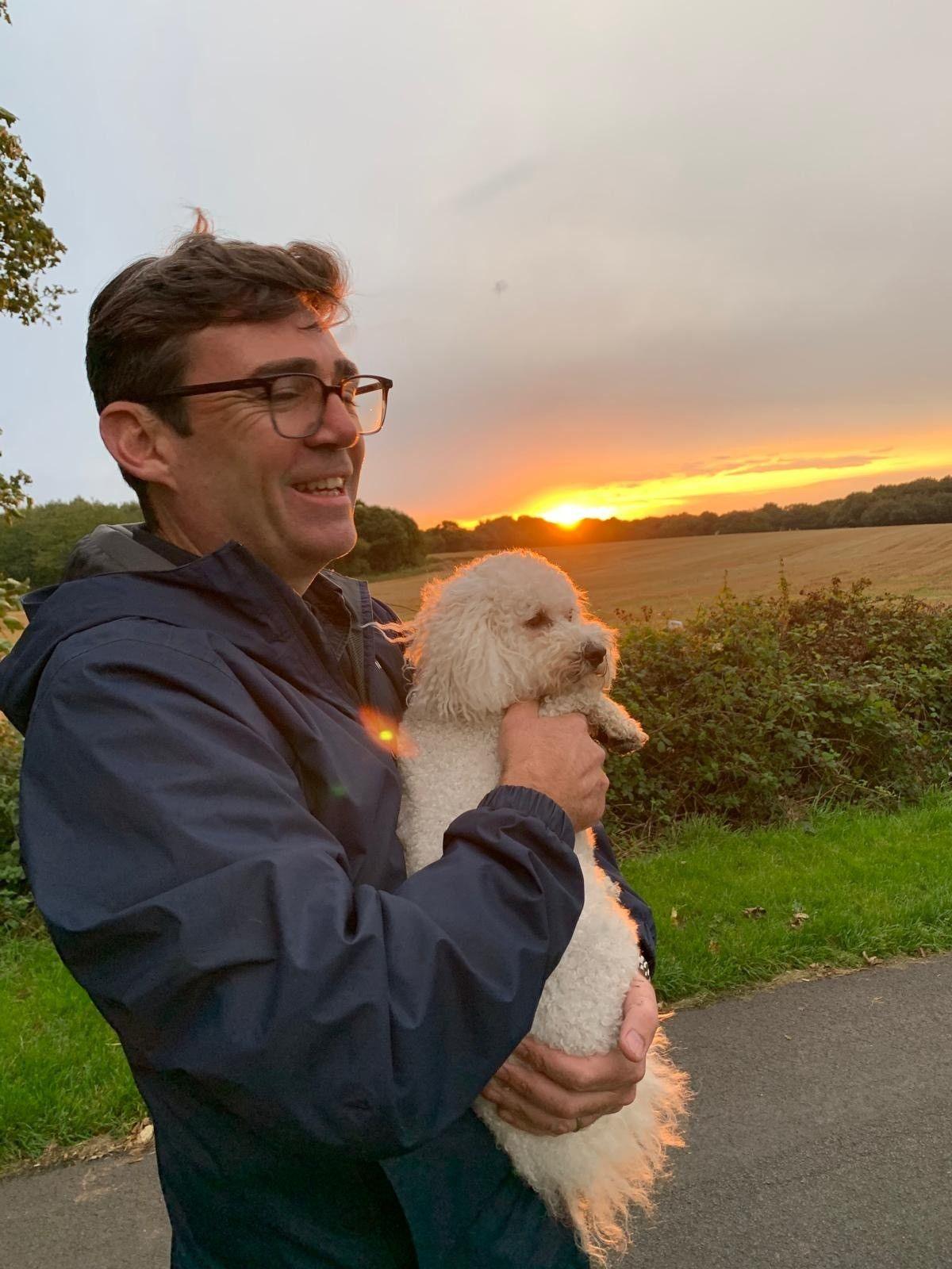 Andy Burnham holding his dog, a white bichon frise, on a country lane with a sunset in the background