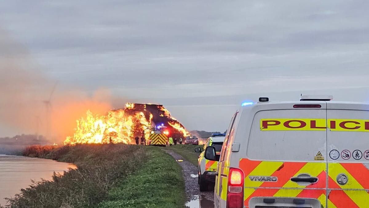 A large fire in the centre of the image surrounded by grass and flat open farm land. The fire is made up of rectangle shaped hay bales which have turned from a light yellow colour to a charred black. Flames can be seen coming out from the stack of hay and dark grey smoke is rising to sky on the left of the image. In the front right of the image is a police van parked behind a police car. Closer to the fire is a fire engine.