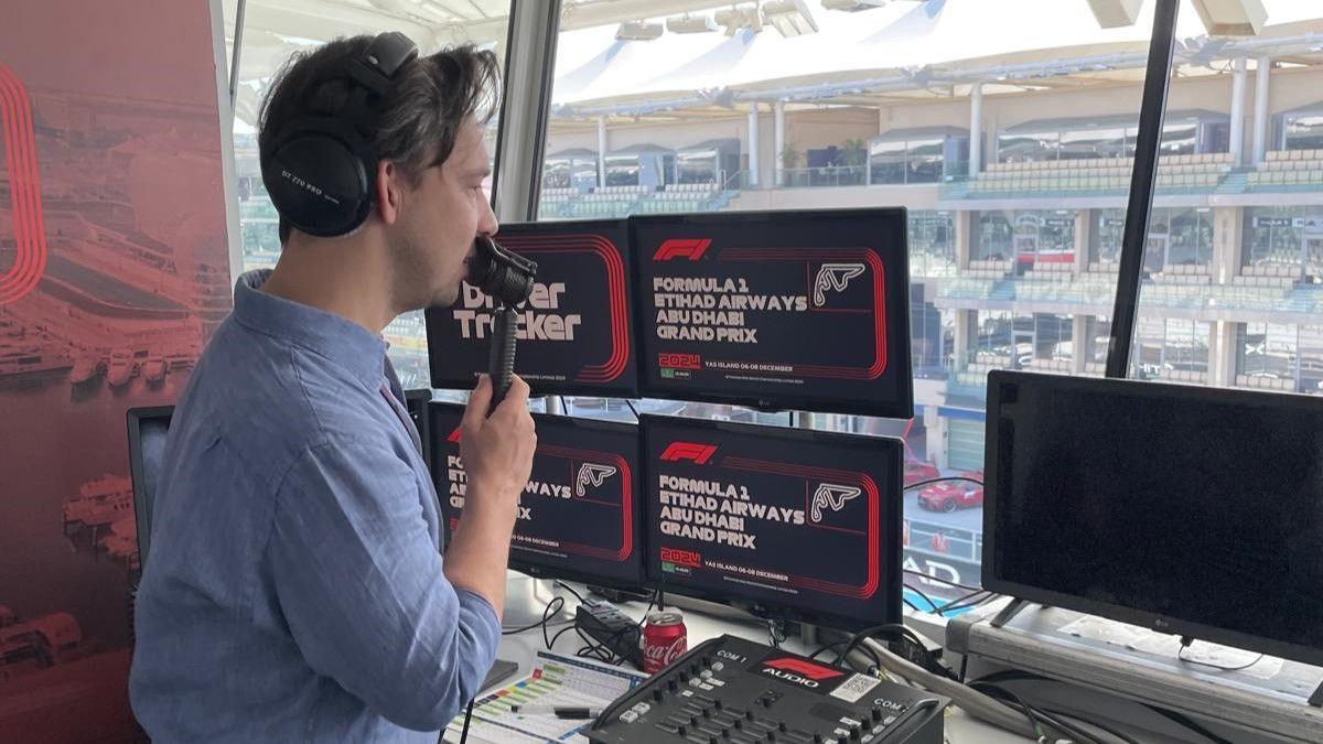 Alex Jacques, side-on the camera, holds a lip mic to his mouth. In front of him are several computer monitors which say "Formula 1 Etihad Airways Abu Dhabi Grand Prix". He is looking out of a window towards a largely empty grandstand.