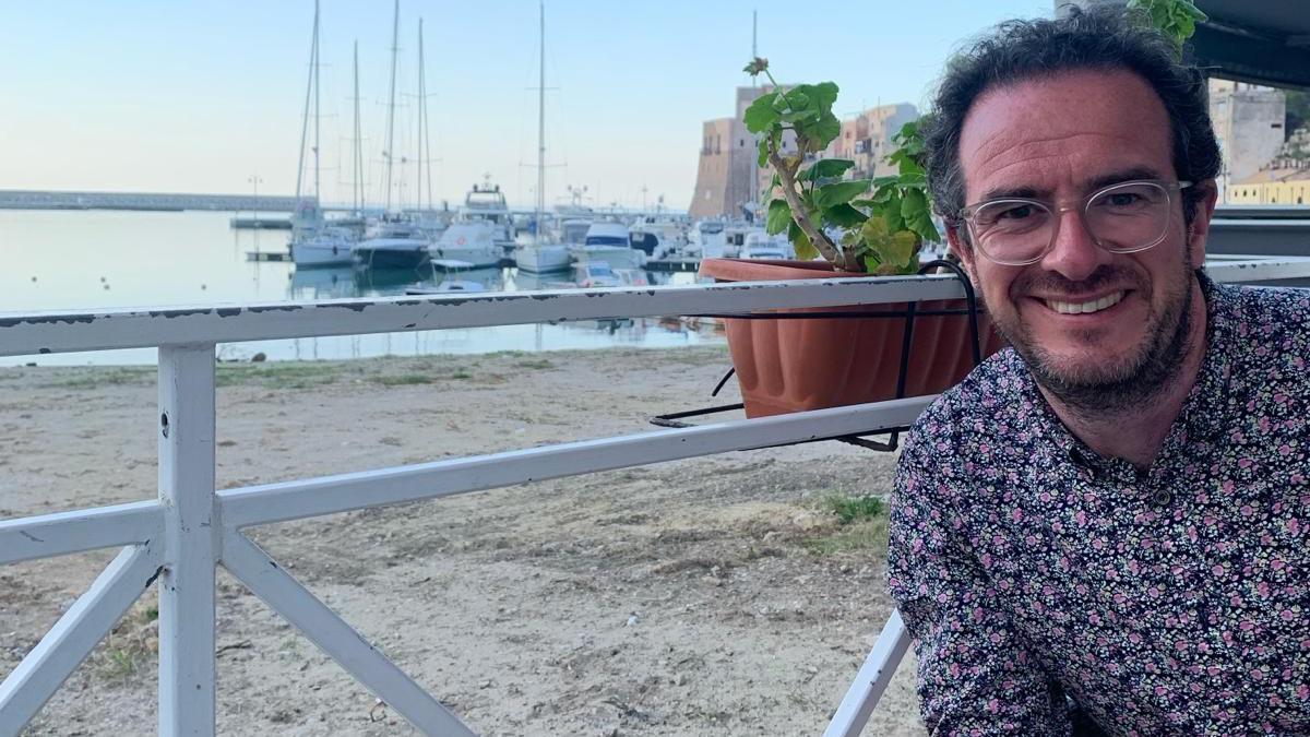 Daniel Allingham sits next to a railing overlooking a port. He wears short hair, glasses and a flowered summery shirt.