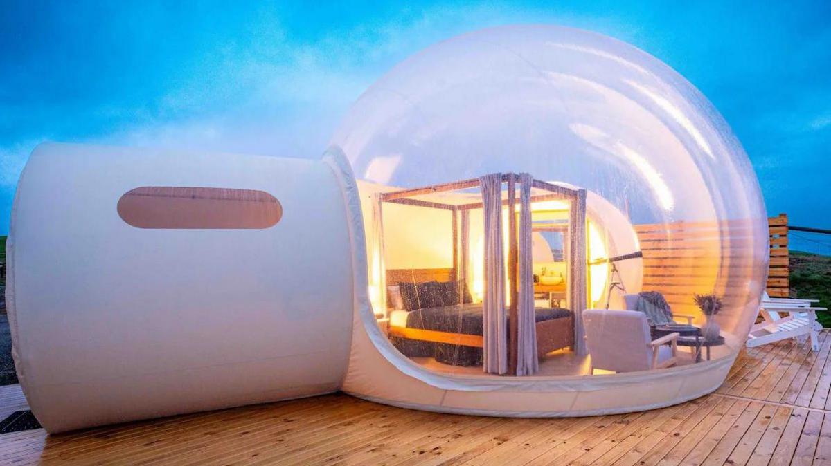 An inflatable white dome with a transparent bubble-like canopy on a wooden deck. There is a four poster bed, a telescope and chairs inside the dome. 