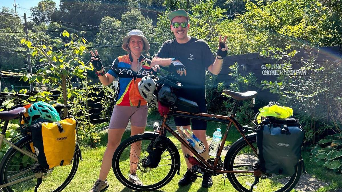 Jess Gray and Matt Kidd stand outside in a garden on a sunny day, with their bikes in front of them.