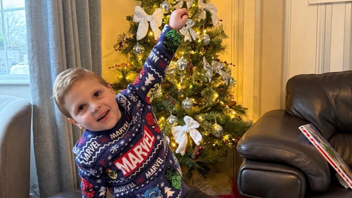 A young boy wearing a Christmas Marvel jumper and smiling with his hand in the air in front of a Christmas tree