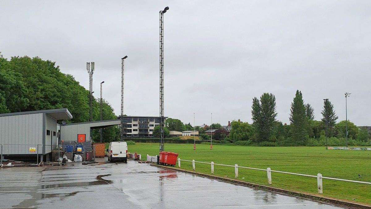 A rugby pitch with car park in the foreground. There is a block of flats in the distance, overlooking the club, and a club house and spectator stand to the left hand side of the image.