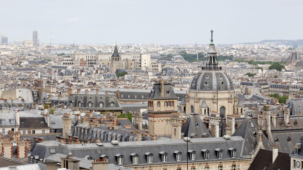 A bird's eye view of Paris in France, the grey roof tops of various buildings can be seen including spire tops, flat roods and dome shaped roofs. Most of the buildings have light stone coloured walls.