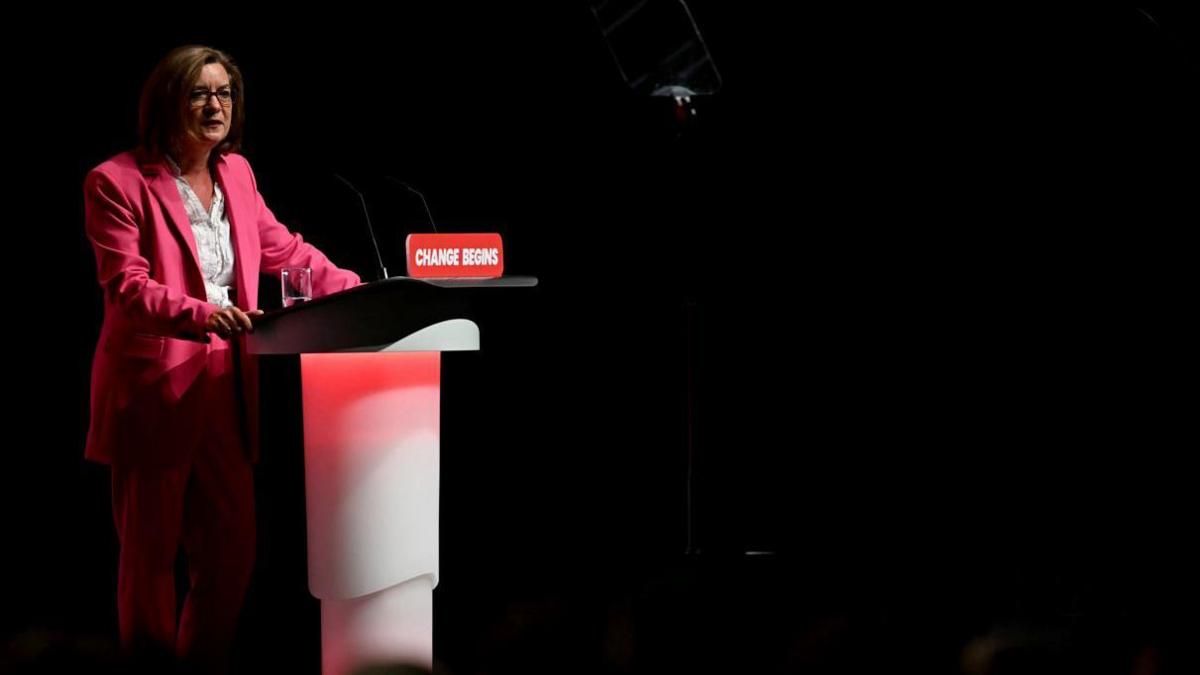 Wales' First Minister Eluned Morgan stood behind a lectern. On it is a sign that says change begins. She is wearing a pink suit with a white shirt.