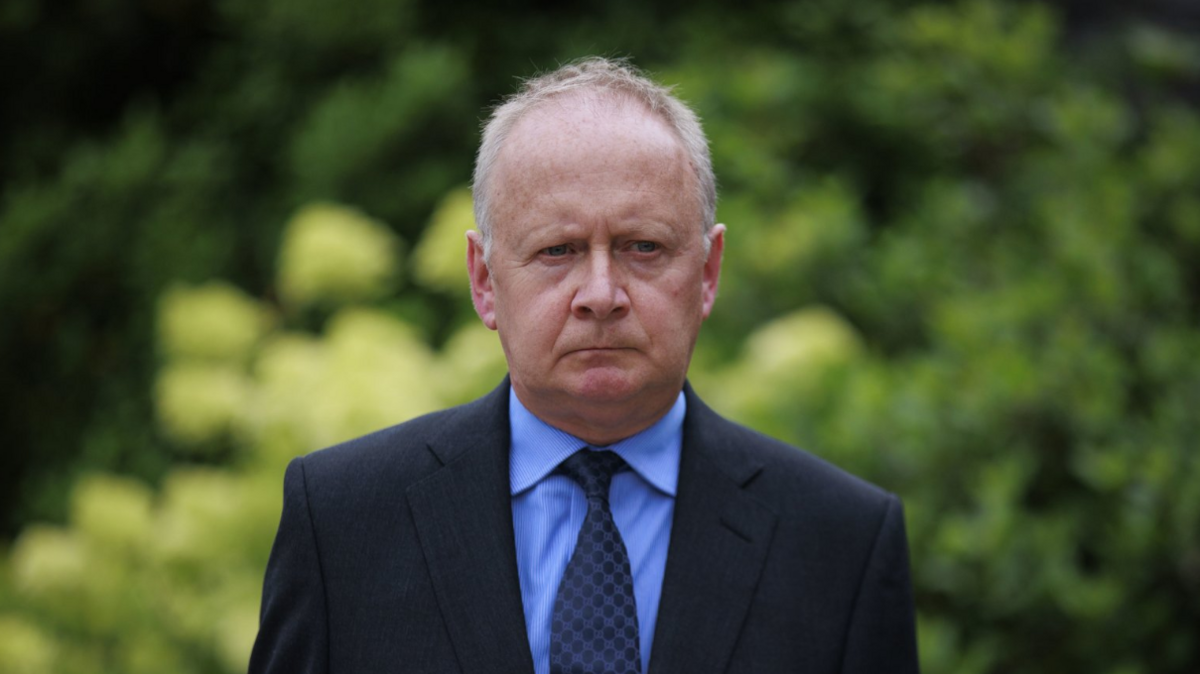 Lord Turnbull, with grey hair, wearing blue jacket, blue shirt and blue tie. In the background - out of focus - is a green hedge.