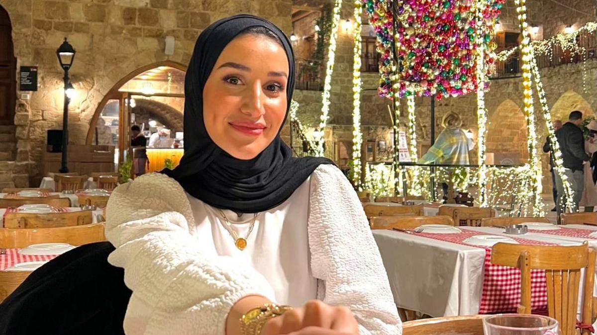 Alaa Ghalayini is sitting in a restaurant covered in festive lighting. She is smiling and wearing a dark headscarf, with a white top and a gold necklace. She appears to be holding the hand of whoever is taking the picture. 