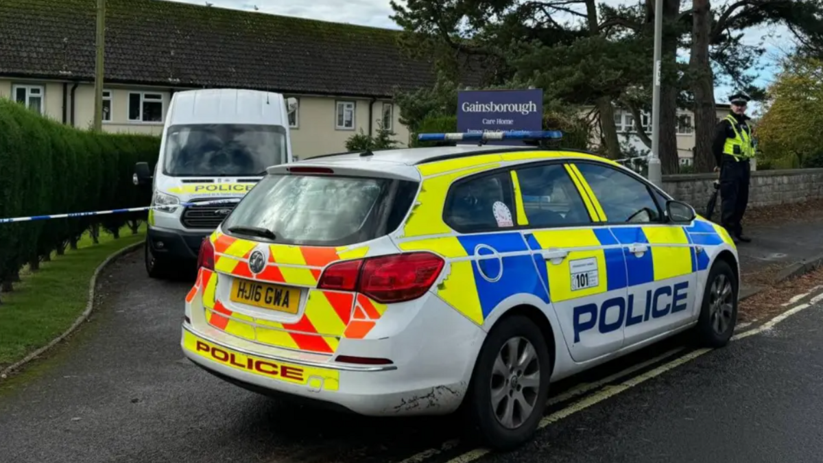 A police car and van are parked either side of police tape at Gainsborough Care Home. A uniformed officer stands guarding the cordon.