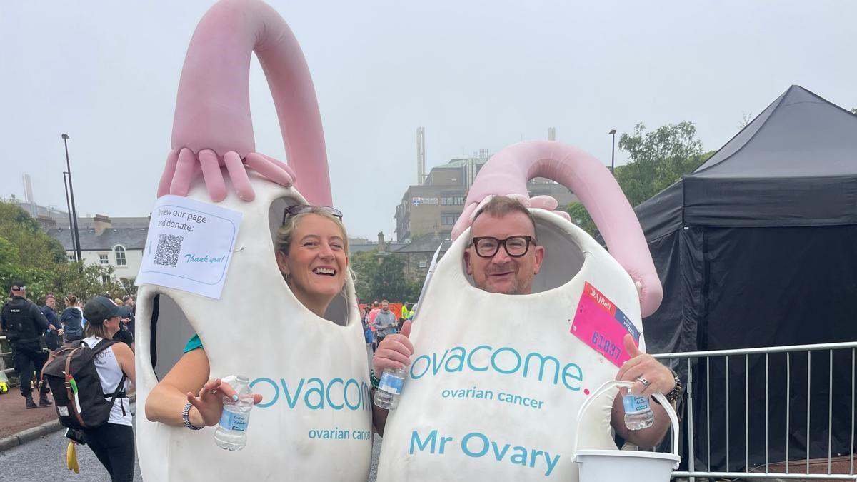 Sarah Temple and Craig McMurough dresses as ovaries at the start of the Great North Run 2024. They are smiling and holding bottles of water. Mr McMurough is also holding a bucket for donations.