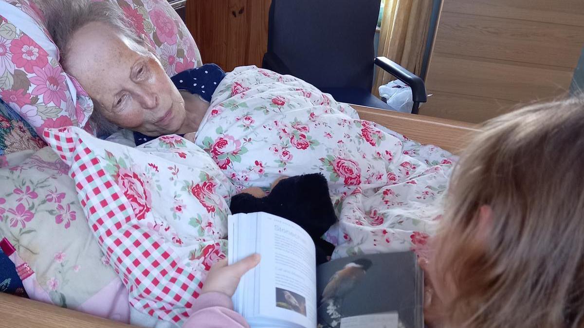 An older woman lies in bed, with floral print duvet. She is wearing a navy top and has grey hair. A child sits beside the bed with brown hair, holding a book with photos of birds.