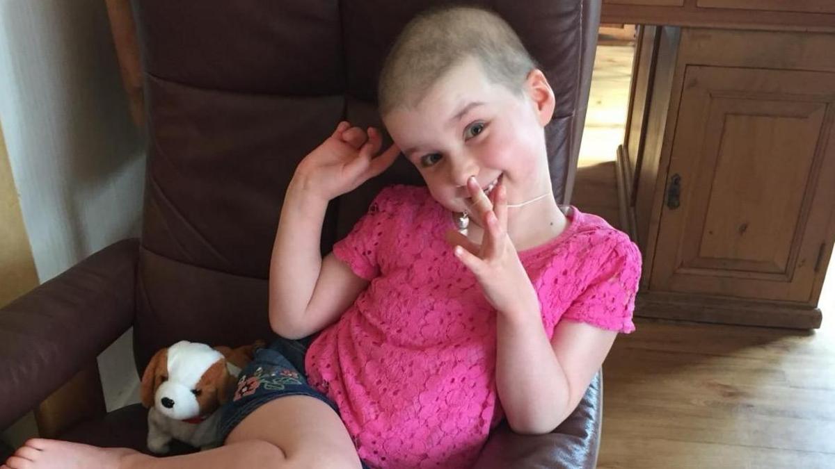 A young girl in a pink dress and shaved hair smiling