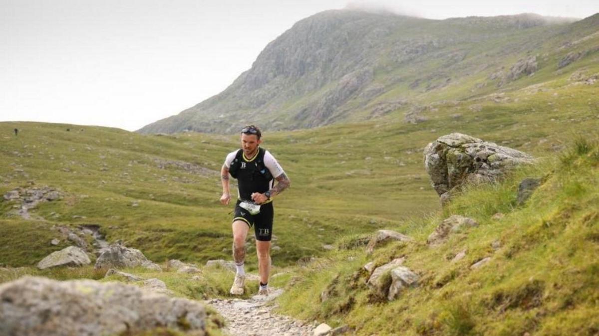 A man running wearing sports gear runs across a large rocky and green open space.