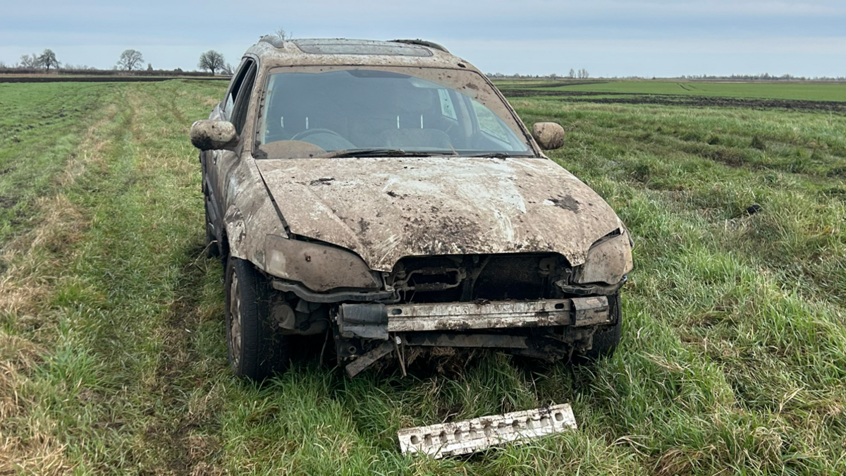 A 4x4 car has been abandoned in a field. It is covered in mud and the front part of the vehicle is hanging off. 