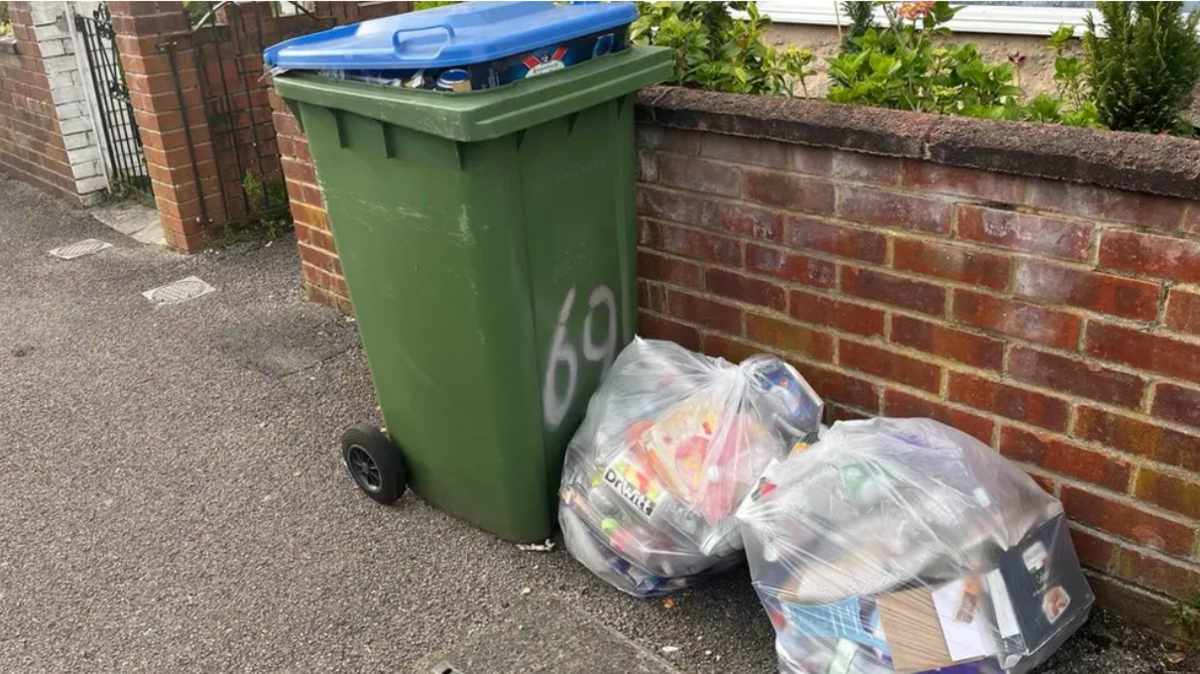 A green Southampton City Council wheelie bin with a blue lid stands on a pavement next to two plastic bags full of waste.