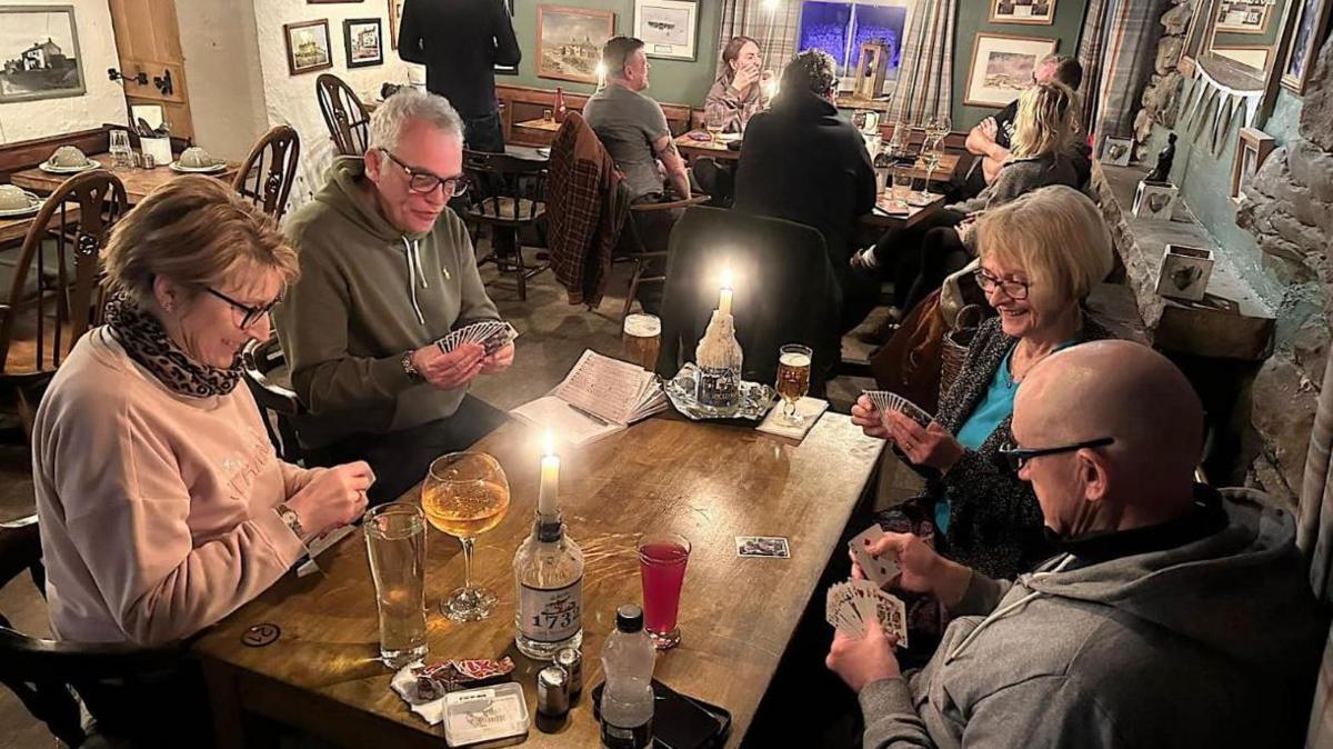 Pub visitors sit around a candle-lit table enjoying drinks and playing cards.