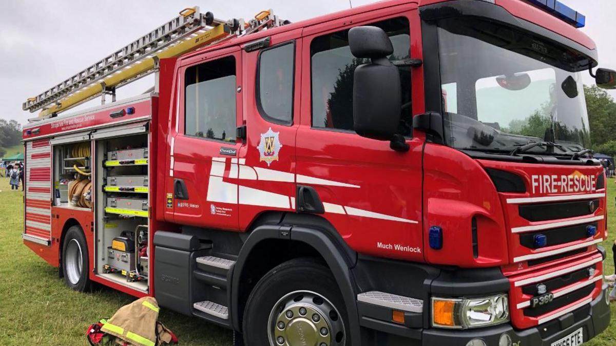 A red fire engine parked on grass with the side panels open and a fireman's light brown jacket on the ground