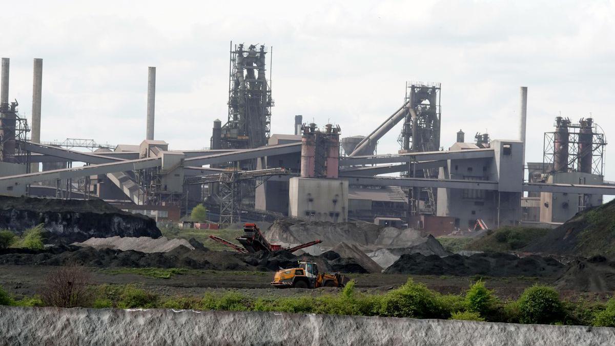Lots of grey buildings and chimneys reach into the sky.  Heaps of material are on the land in the foreground and red and yellow machinery is working next to them.