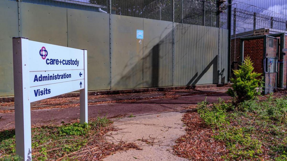 A signpost reads "CARE+CUSTODY" with arrows that show the way to the visiting and administration centre inside the abandoned facility of the Immigration Removal Centre Campsfield in Kidlington near Oxford. Its walls are fenced with barb wire.