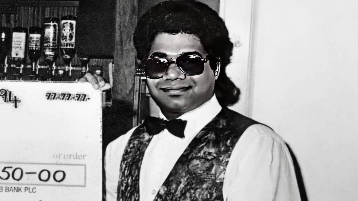 Black and white photograph of Shamsuddin Mahmood in a restaurant holding a big cheque. He is smiling. He has long, dark, swept-back hair. He is wearing a white shirt, black bow tie and a patterned waistcoat.