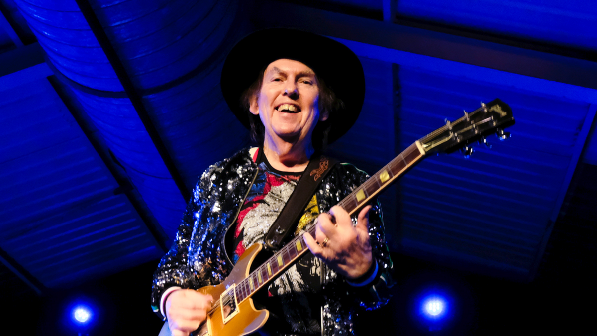 A man wearing a cowboy hat and sparkly jacket on stage with a brown wooden-panelled guitar