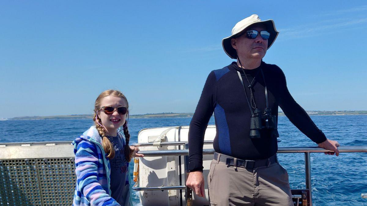 A young girl stood next to a middle aged man. They are both wearing sunglasses and are on a boat. The sky behind them is blue, with the horizon in the distance.