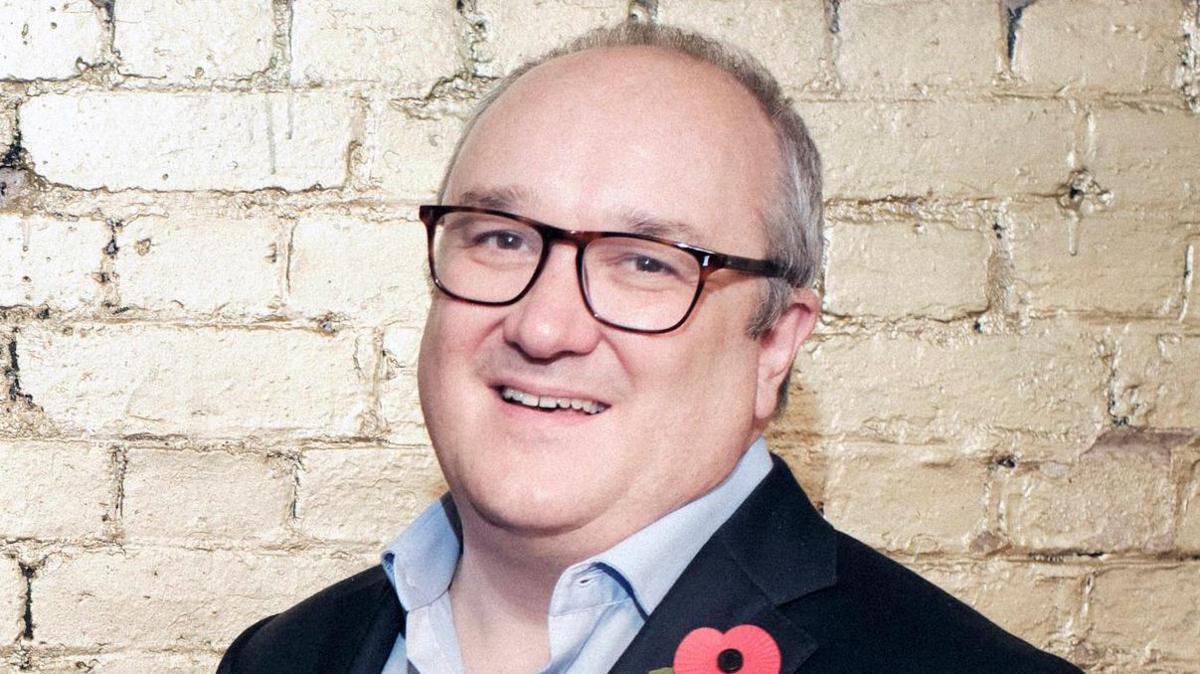 Dominic Sandbrook has extremely receding dark and grey hair. He is wearing black-rimmed square spectacles on his face and a navy suit blazer with a red poppy on the lapel over a blue shirt, which is unbuttoned at the collar. He is smiling at the camera.