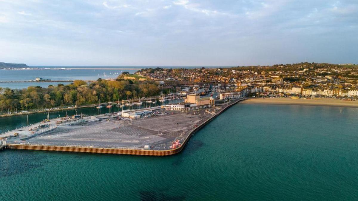 Aerial view, looking from the sea towards Weymouth Peninsula and Weymouth Beach. The peninsula is a flat triangle of land, mostly covered by a large car park. To the right of the peninsula is the sandy beach. To the left is the entrance to the harbour which is full of small boats on their moorings. On the far side of the harbour entrance is the tree-filled peninsula of Nothe Gardens.