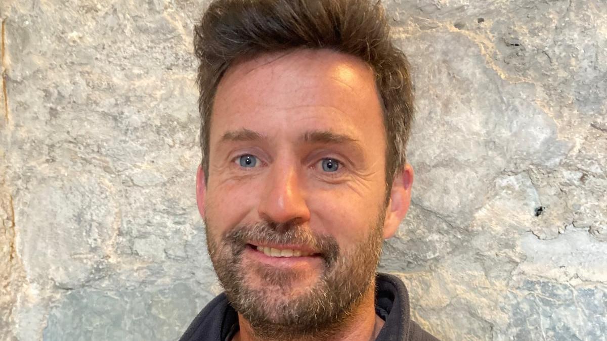 Ryan Byrne has blue eyes, brown hair and a beard.
He is wearing a grey fleece and standing in front of a stone wall. 