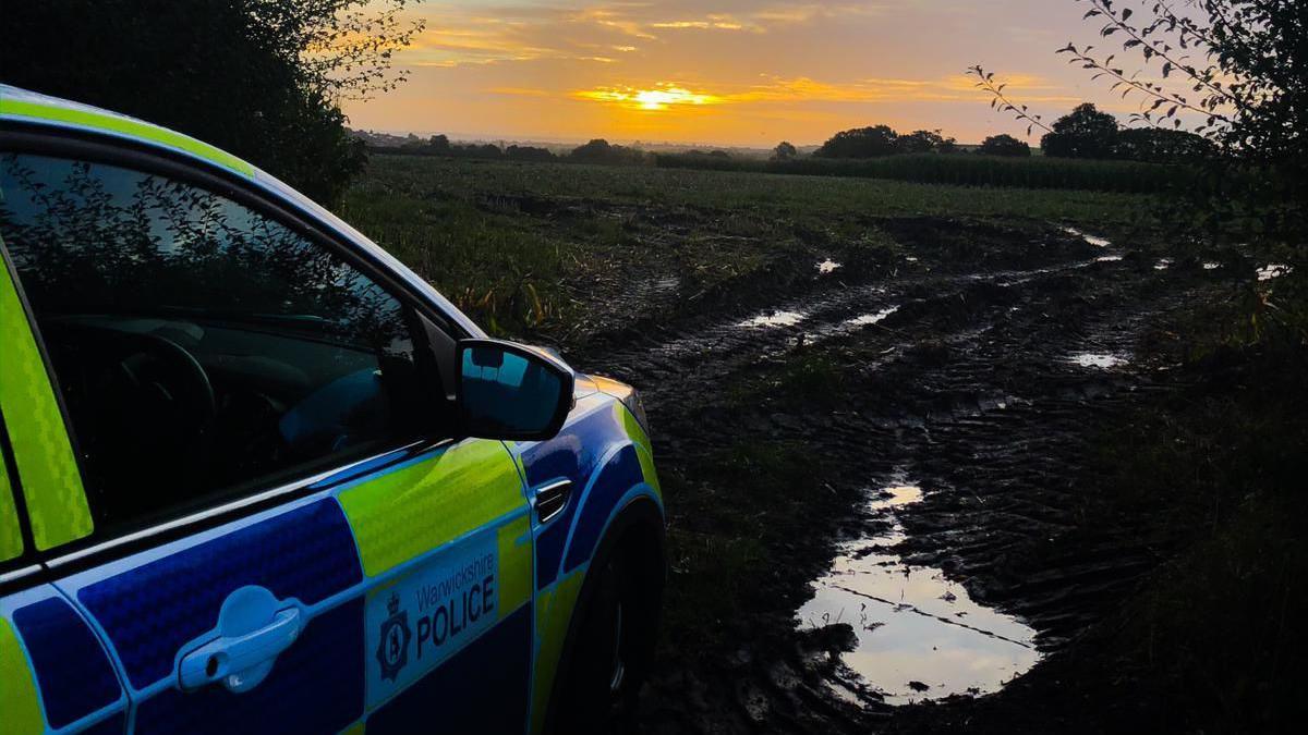 A Warwickshire Police vehicle on muddy field. It is parked off to the side and the sun is low in the sky during sunset. 