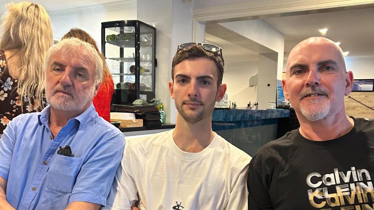 Three men sat in a public venue of some description. The one on the left is in his seventies, has white hair and a beard and is wearing a blue shirt. The man in the middle is in his twenties and wearing a white t-shirt.. The man on the right is in his fifties and wearing a black t-shirt.