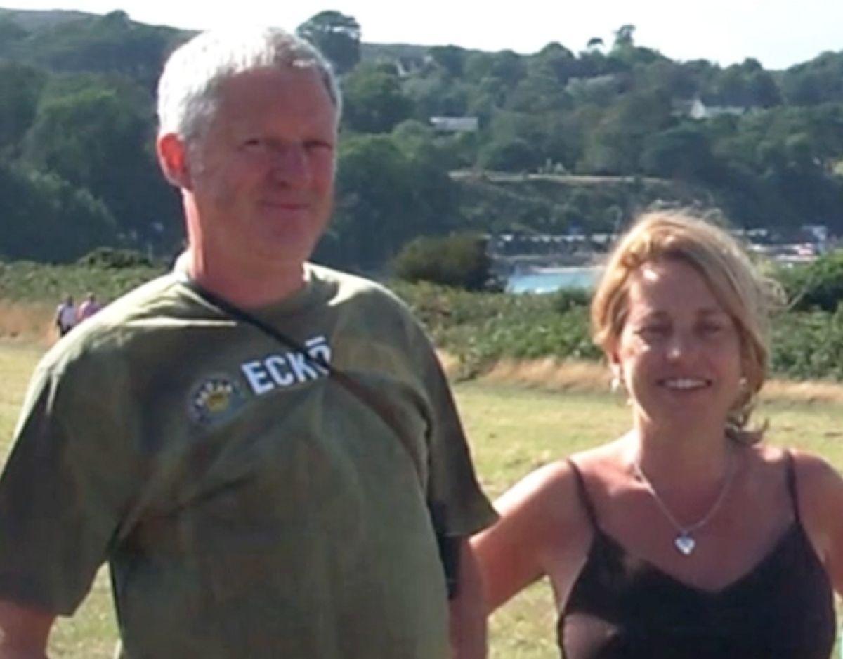 A man and a woman are pictured in the countryside by an expanse of water. It is a sunny summer's day and trees are in full leaf on the hillside in the distance. The man is wearing a green T-shirt, he has straight grey hair and is smiling at the camera. The woman is also smiling. She has straight blond hair, is wearing a necklace with silver heart, and is wearing a brown summery top with thin shoulder straps.