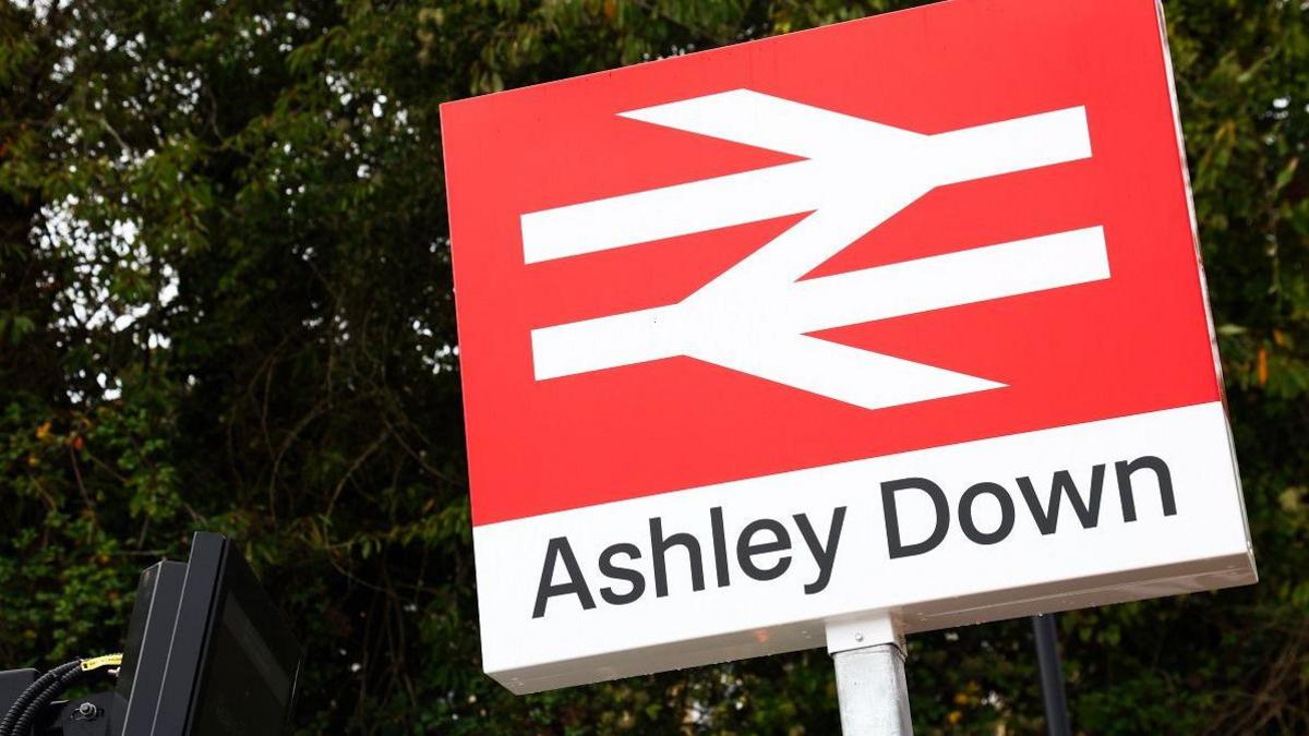 A red sign with a white train line logo and the words Ashley Down written in black with trees in the background