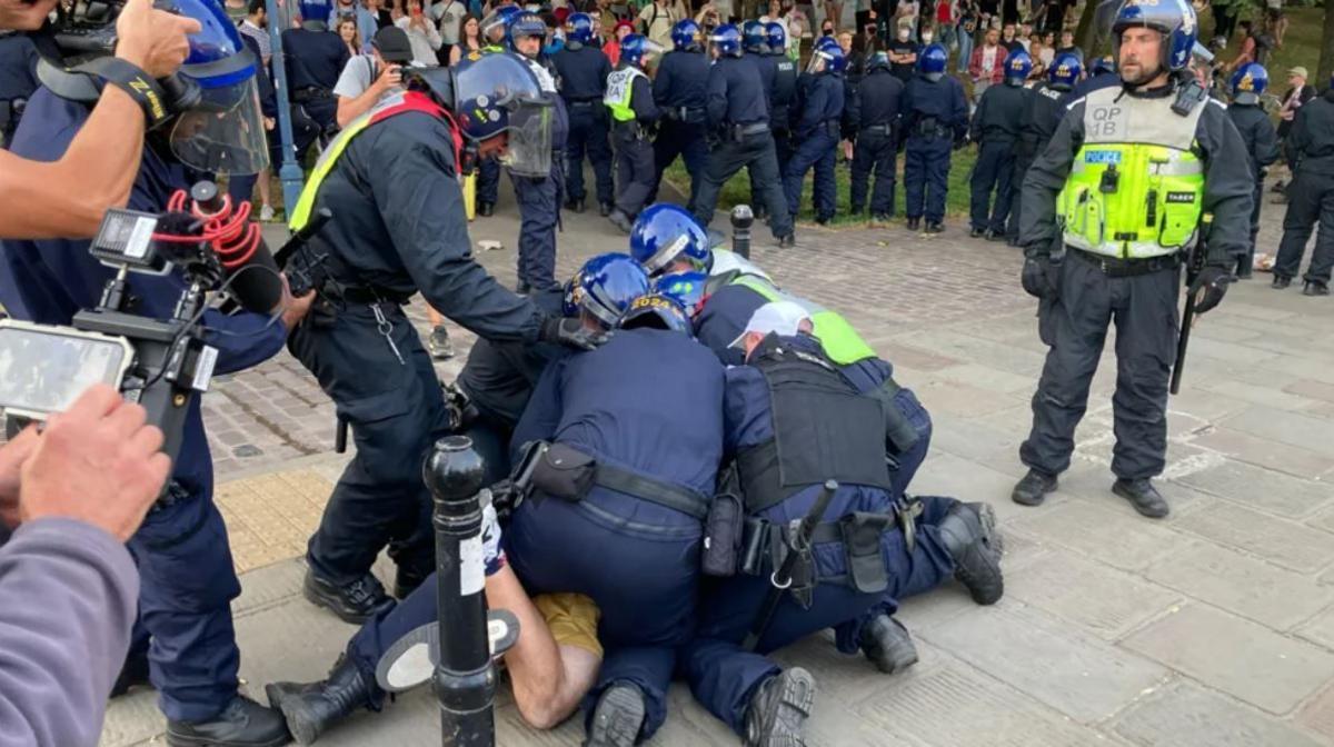 Five police officers kneeling on top of a suspect while other police officers try to keep protesters at bay