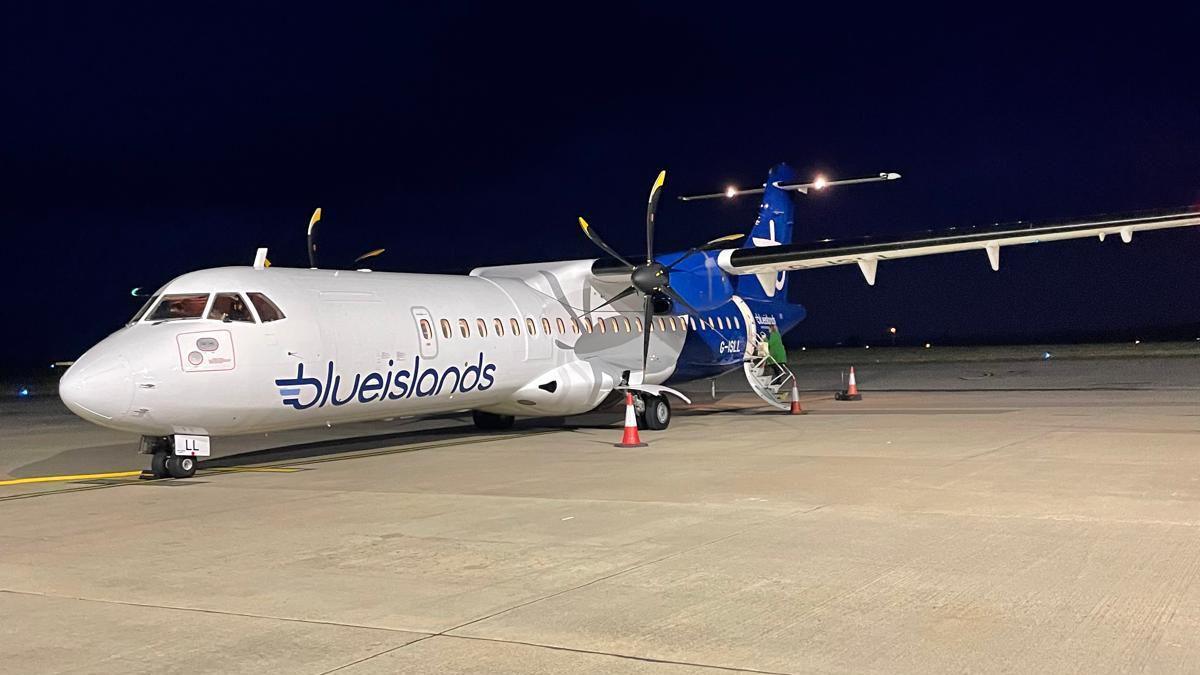 Blue Islands plane, white at the front and dark blue at the back with two propellers, on a beige runway late at night