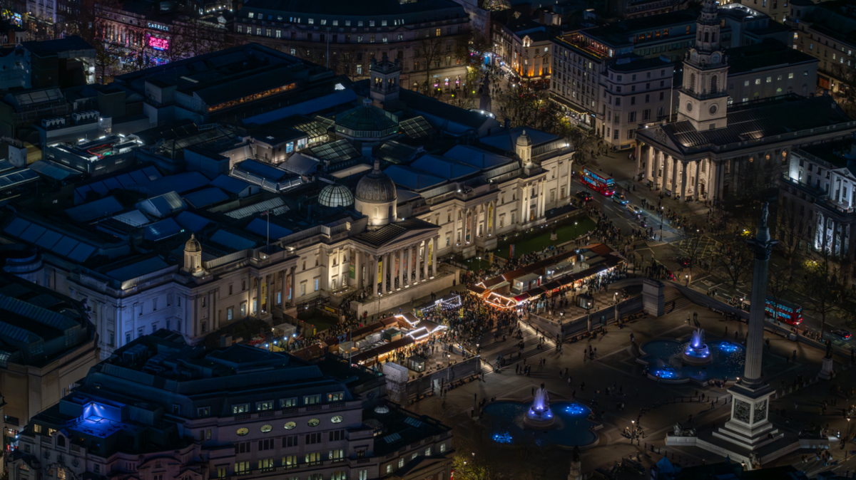 Aerial photograph taken at night showing Trafalgar Square with various huts of the Christmas market lit up and surrounded by people with the National Gallery behind them