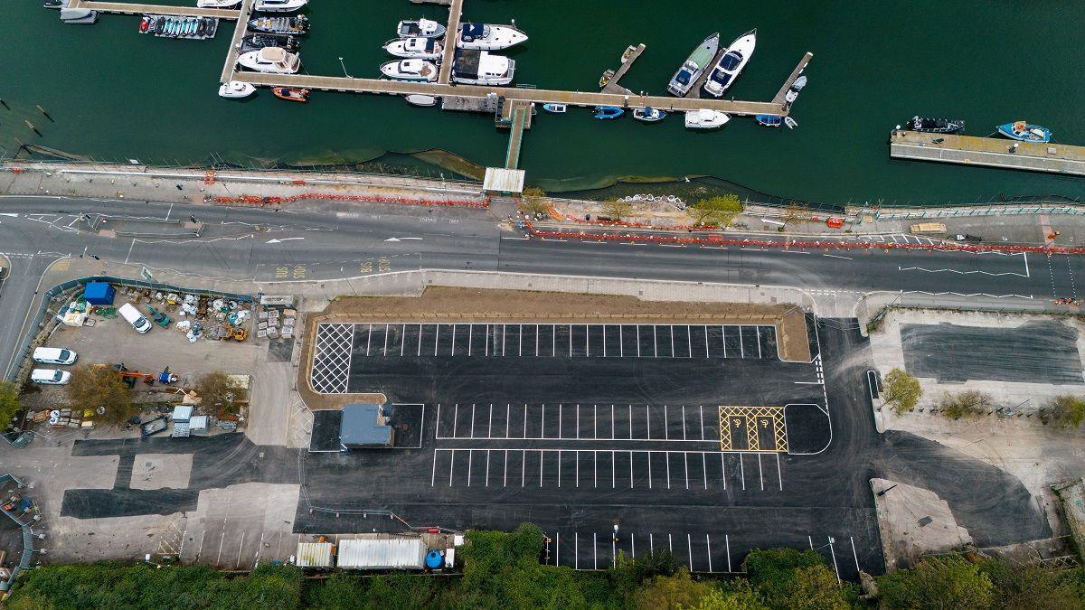 A drone photo of a car park from above. It's empty and sits on what appears to be an industrial site next to a waterfront, with a T-shaped pontoon that has boats moored up along it.