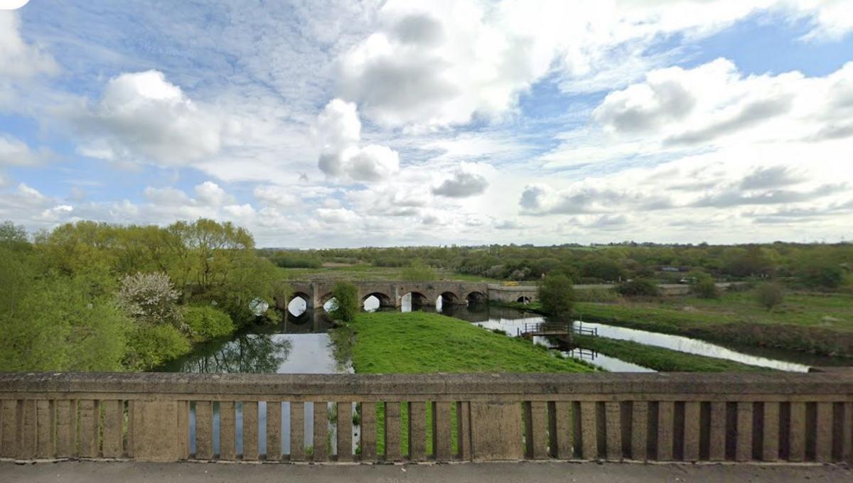 Image of Stanwick Lakes showing a low stone parapet with narrow lakes behind, separated by a grass area. An arched bridge is visible in the distance.