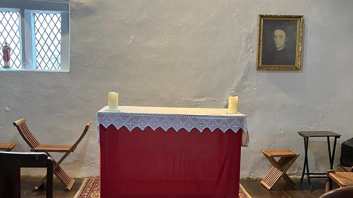 The altar in the attic in Brindle where Edmund Arrowsmith celebrated his last Mass. It has a white lace cover and a red cloth draped down to the floor. There are two candles on it, and a painting of the saint hangs on the wall.