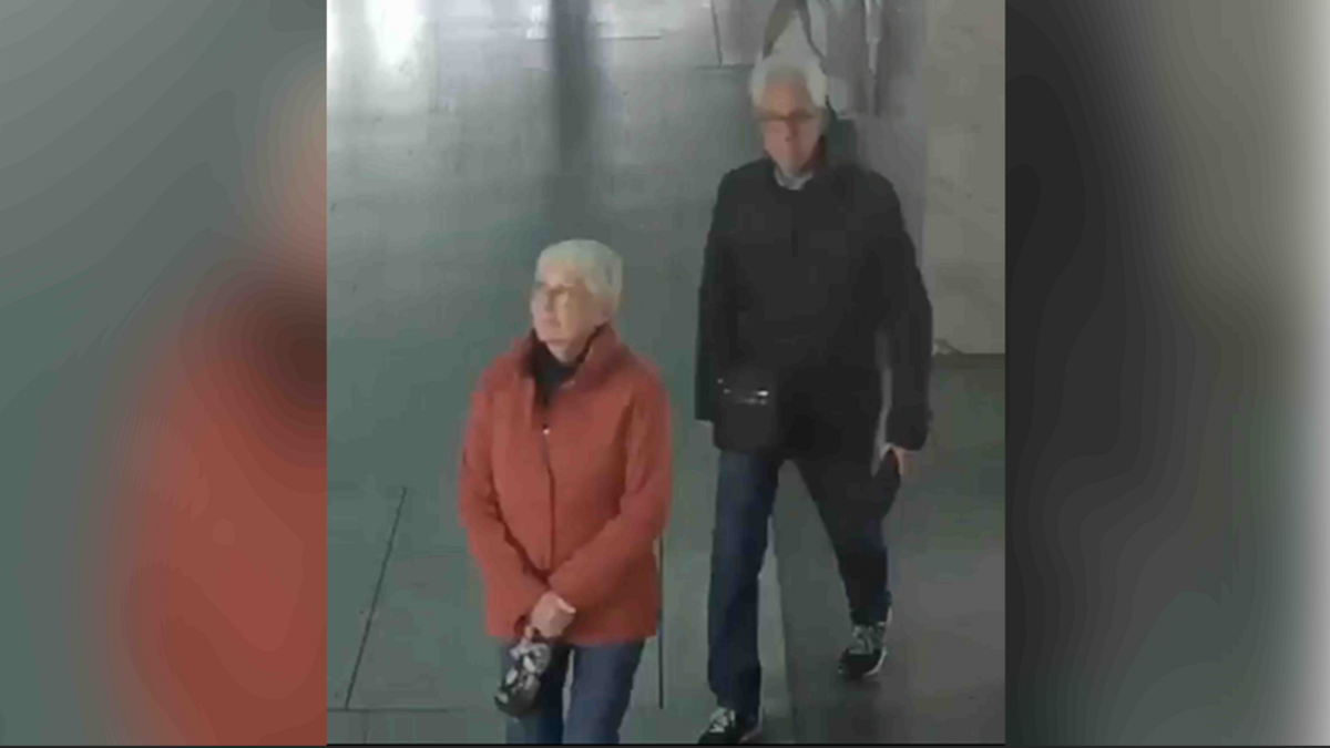 Woman in orange jacket and man in black jacket, both wearing jeans and both with grey hair. They are walking across a paved or tiled floor.