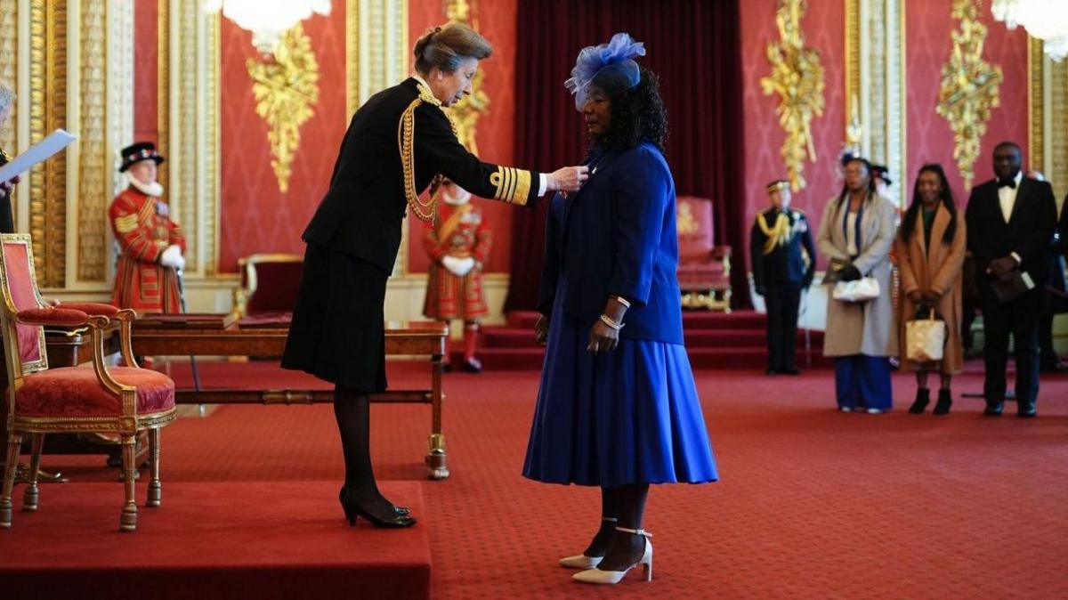 Dr Gee Walker stands in front of the Princess Royal who is placing the MBE on her blue blazer. Her family are watching in the background. The room is adorned with red and gold. 